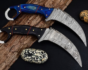 Può includere: Due coltelli curvi con lame in acciaio Damasco e manici in legno. Il coltello superiore ha un manico blu con dettagli dorati, e il coltello inferiore ha un manico nero con dettagli dorati.