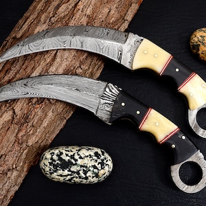 May include: Two curved, black-handled knives with white and red accents. The blades are made of Damascus steel and have a distinctive wavy pattern. The knives are displayed on a piece of wood with a rough texture.