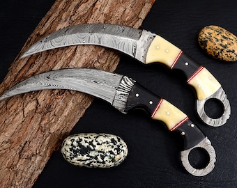 Può includere: Due coltelli curvi con manici neri, dettagli bianchi e rossi. Le lame sono in acciaio Damasco e presentano un motivo ondulato distintivo. I coltelli sono esposti su un pezzo di legno con una texture ruvida.