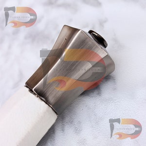 May include: Close-up of the top of a sword hilt. The hilt is made of metal and has a square shape. The metal has a brushed finish and is a silver color. The top of the hilt is capped with a round, black piece of metal.