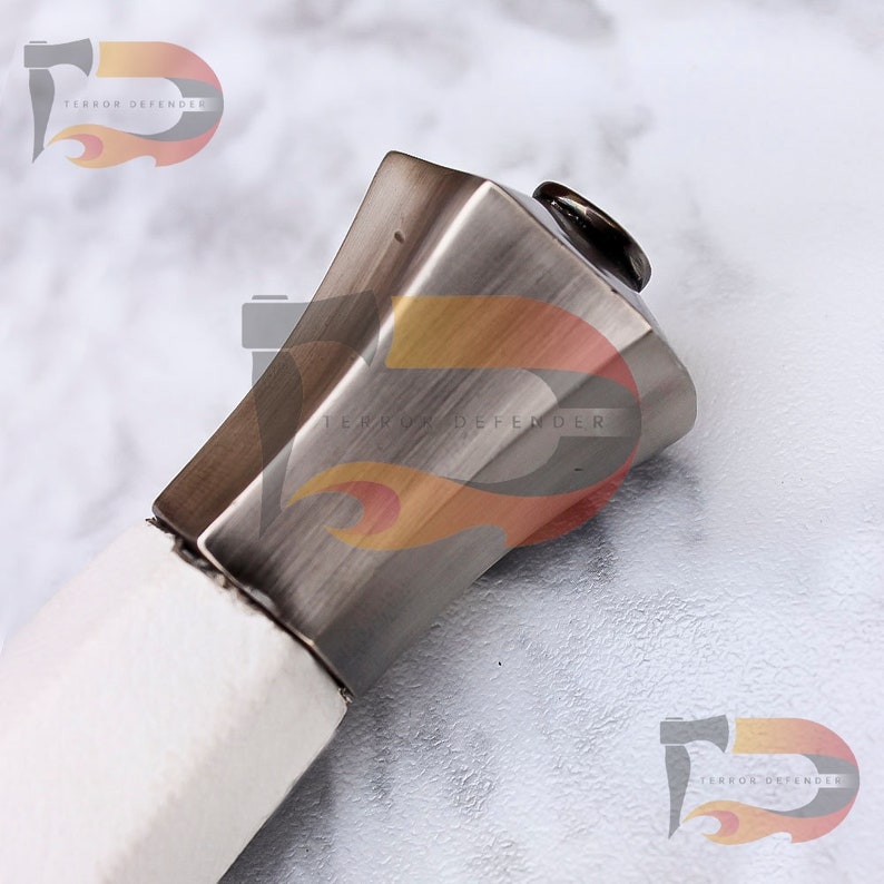 May include: Close-up of the top of a sword hilt. The hilt is made of metal and has a square shape. The metal has a brushed finish and is a silver color. The top of the hilt is capped with a round, black piece of metal.