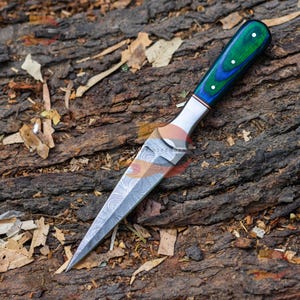 May include: A Damascus steel dagger with a green and blue wooden handle. The blade has a detailed pattern, and the handle features three white dots. The dagger is displayed on a textured, brown wooden surface.