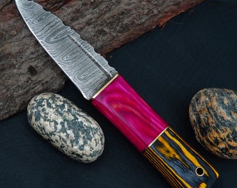Può includere: Un coltello in acciaio Damasco con manico in legno rosa e nero. La lama presenta un motivo ondulato e il manico è decorato con un accento dorato.
