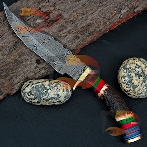 Könnte beinhalten: Ein Jagdmesser mit einer Damaststahlklinge und einem Hirschhorn-Griff. Der Griff ist mit roter, grüner und blauer Schnur umwickelt. Das Messer liegt auf einem Stück Holz und zwei Steinen.
