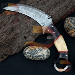 May include: A curved, silver-coloured knife with a black and white handle. The handle has a red accent and a ring at the end. The knife is resting on a piece of wood and a rock.