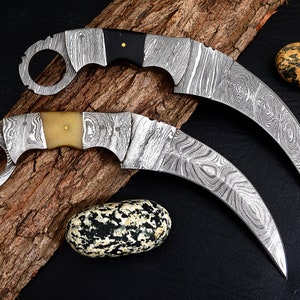 May include: Two curved knives with a ring handle and a Damascus steel blade. The knives have different colored handles, one is black and the other is white.