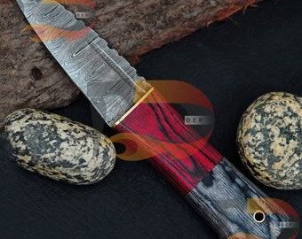 Può includere: Un coltello in acciaio Damasco con manico in legno rosso e nero. La lama presenta un motivo ondulato e un accento dorato vicino al manico.