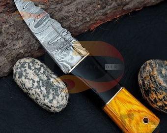Può includere: Un coltello in acciaio Damasco con manico in legno nero e giallo. La lama presenta un motivo ondulato e un bordo seghettato. Il manico è decorato con un accento in ottone.