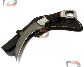 Può includere: Un coltello ricurvo color argento con manico nero e fodero in pelle. Il coltello ha un motivo decorativo sulla lama e un anello sul manico.