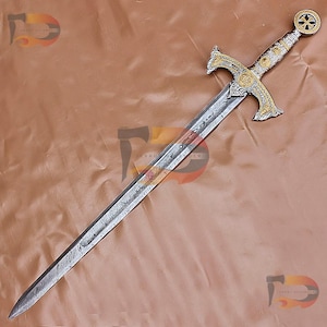 May include: A silver and gold sword with a decorative handle and a patterned blade. The sword is on a brown background.
