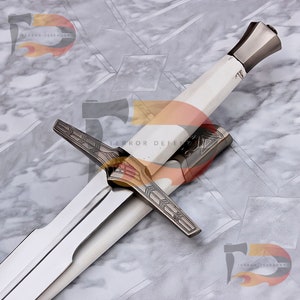 May include: A silver and gold sword with a white handle and intricate details. The sword has a cross guard with a decorative design. The sword is on a gray marble surface.