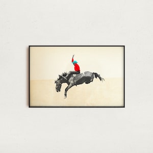 May include: A framed art print featuring a cowboy riding a bucking bronco. The cowboy is wearing a red shirt and a blue hat. The horse is black and white, and the background is a light beige color.