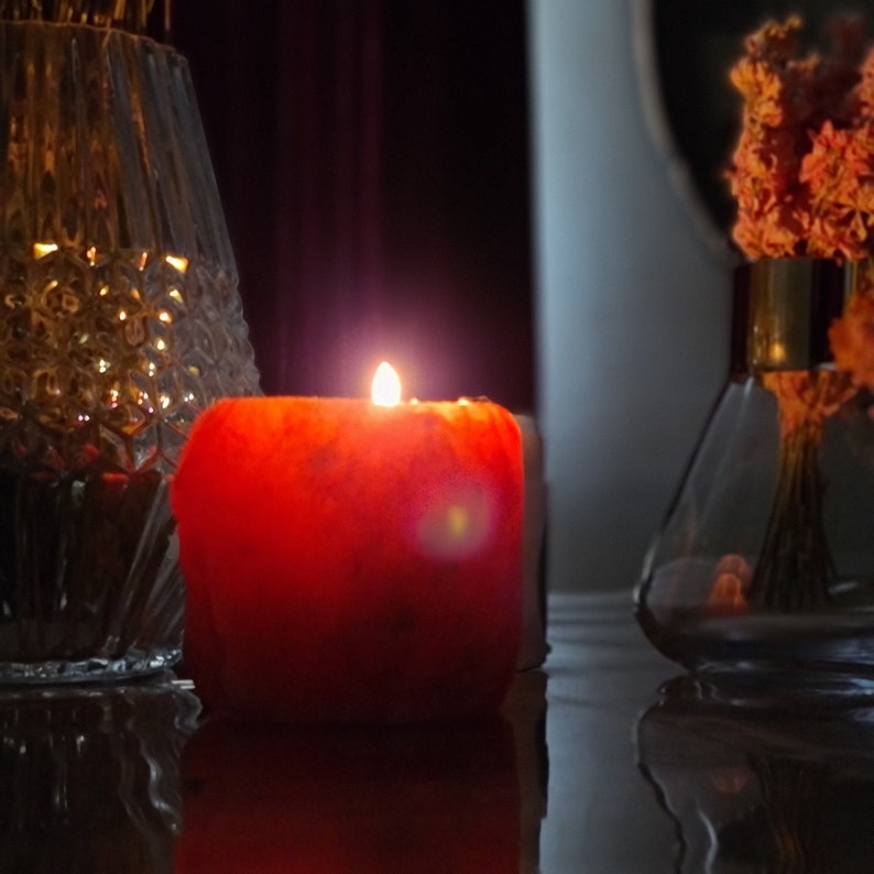 May include: A red candle with a warm glow sits on a table next to a clear glass vase filled with dried flowers.