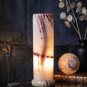 Puede incluir: Lámpara cilíndrica de ónix con un patrón veteado crema y morado. La lámpara está iluminada desde dentro, proyectando un brillo cálido. Una esfera a juego y un jarrón negro con follaje seco están en el fondo.