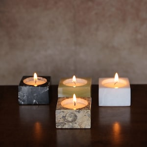 May include: Four lit candles in different colored marble candle holders. The candles are in square holders, one black, one white, one green, and one brown with white flecks.