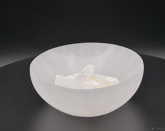 May include: A white selenite bowl filled with selenite sticks. The bowl is smooth and has a slightly rounded shape. The selenite sticks are white and have a natural, rough texture.