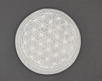 Peut inclure: Une plaque ronde en cristal de sélénite blanche, ornée d'un motif de Fleur de Vie gravé. Le motif est mis en valeur sur un fond gris foncé, soulignant les détails.