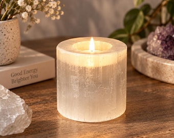 May include: A cylindrical selenite crystal candle holder with a translucent, fibrous white texture. The stone is hollowed at the top to hold a small, lit candle that emits a warm, soft glow, illuminating the natural vertical striations of the mineral.