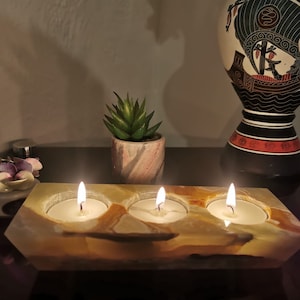 May include: A three-wick candle holder made of natural onyx stone. The holder has three circular depressions for tealight candles. The candles are lit and glowing. The holder is on a dark surface with a small potted succulent plant and a decorative vase in the background.