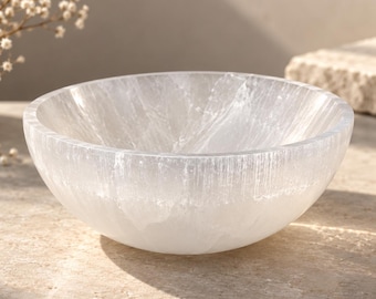 May include: A round, translucent white selenite bowl with a smooth, polished interior and a fibrous, natural crystalline texture on the exterior. The bowl features a bright, reflective surface that catches light, showcasing the mineral's unique layered structure and milky, semi-transparent appearance.
