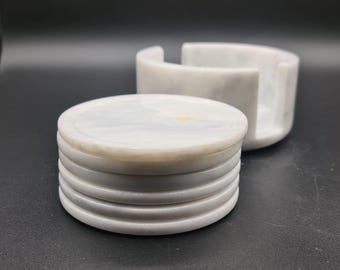May include: A stack of five circular white marble coasters with subtle grey veining and a smooth finish, accompanied by a matching cylindrical marble holder with a side cutout, designed to store the coasters when they are not in use.