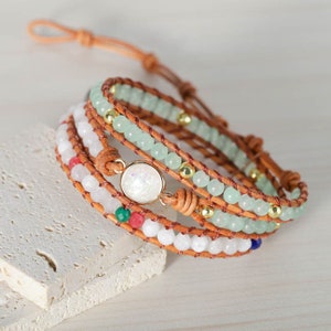 May include: A multi-strand bracelet with brown leather cord, white, green, and red beads, and a gold-toned metal accent. The bracelet features a large, white, iridescent stone.