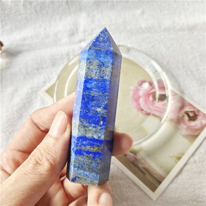 May include: A deep blue lapis lazuli crystal point with gold flecks. The crystal is faceted and has a pointed tip. It is held in a hand.