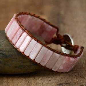 May include: A pink gemstone bracelet with a brown leather strap and a silver clasp. The bracelet is made of rectangular shaped stones.