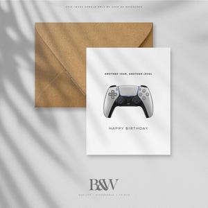 May include: A white greeting card with a gray and black game controller illustration. The card reads "ANOTHER YEAR, ANOTHER LEVEL" and "HAPPY BIRTHDAY". A brown envelope is behind the card.