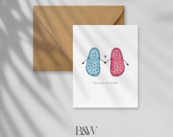 May include: A greeting card with a brown envelope. The card features two cartoon Crocs, one blue and one pink, holding hands with a small heart between them. The text "You croc my world" is below the Crocs.