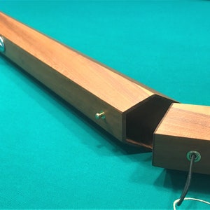 May include: A wooden case for a pool cue, open to reveal the interior. The case is made of light brown wood with a dark brown stain. The case has a hexagonal shape and is held together with a metal pin.