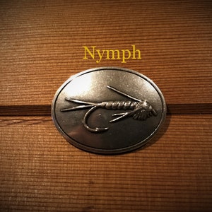 May include: A silver oval-shaped pin with a detailed image of a fly fishing lure. The pin has the word "Nymph" printed above it.