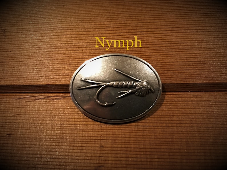 May include: A silver oval-shaped pin with a detailed image of a fly fishing lure. The pin has the word "Nymph" printed above it.