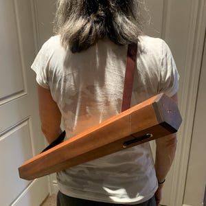 May include: A person wearing a white t-shirt and black shorts is carrying a long, rectangular wooden case with a brown leather strap over their shoulder.