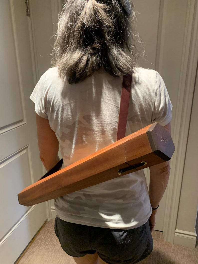May include: A person wearing a white t-shirt and black shorts is carrying a long, rectangular wooden case with a brown leather strap over their shoulder.