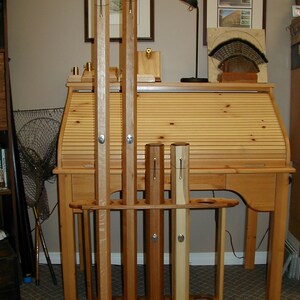 May include: A wooden rod holder with five wooden rods standing upright. The holder is made of light brown wood and has a dark brown stain. The rods are different lengths and have a natural wood finish.