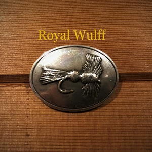 May include: A silver oval-shaped pin with a detailed image of a Royal Wulff fly.