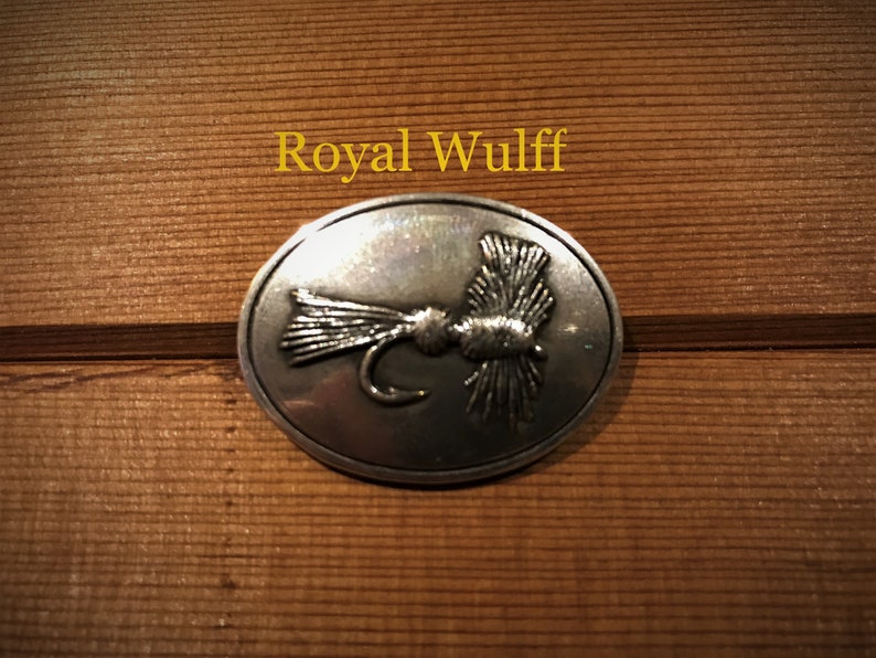 May include: A silver oval-shaped pin with a detailed image of a Royal Wulff fly.