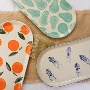 Könnte beinhalten: Drei dekorative Keramik-Servierplatten mit handgemalten Designs. Eine zeigt Orangen, eine andere blaue abstrakte Formen und die dritte blaue Quallen. Die Platten befinden sich auf einer beigefarbenen Jutefläche.