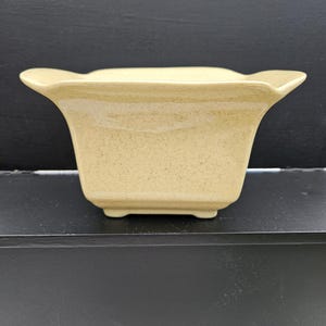 May include: A beige ceramic planter with a flared, scalloped rim. The square-shaped planter has a speckled texture and sits on small feet. The neutral color and unique shape make it a decorative item for plants or other small items.