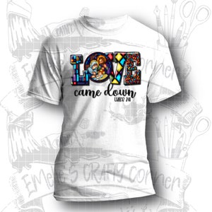 Puede incluir: Camiseta blanca con un dise&ntilde;o de vitral colorido de la palabra "LOVE" con una representaci&oacute;n de la Sagrada Familia dentro de la letra "O". El texto "came down" y "LUKE 2:11" est&aacute;n debajo del dise&ntilde;o.