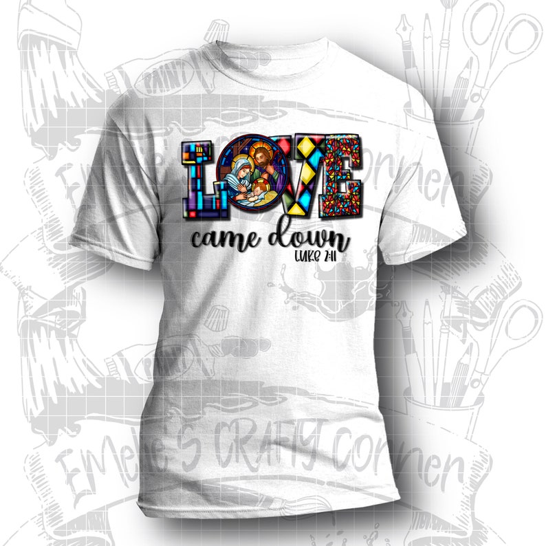 Puede incluir: Camiseta blanca con un dise&ntilde;o de vitral colorido de la palabra "LOVE" con una representaci&oacute;n de la Sagrada Familia dentro de la letra "O". El texto "came down" y "LUKE 2:11" est&aacute;n debajo del dise&ntilde;o.