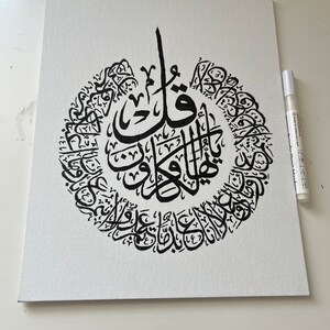 May include: A black and white canvas painting with Arabic calligraphy in a circular design. The calligraphy is in a traditional style and is centered on the canvas.