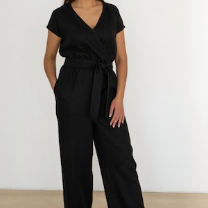 Woman wearing a black linen jumpsuit with short sleeves, V-neck wrap front, waist tie, and wide-leg pants – minimalist, elegant outfit suitable for casual or semi-formal occasions.