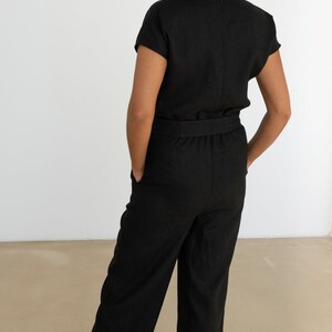 Back view of a woman wearing a black linen jumpsuit with short sleeves, tailored waistband, and wide-leg pants, styled with black heels – elegant and minimalist outfit for various occasions.