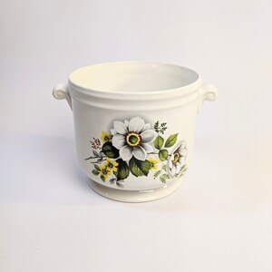 May include: White ceramic planter with two handles and a floral design featuring white, yellow, and green flowers.