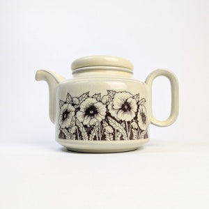 May include: Cream-coloured ceramic teapot with a matching lid. The teapot features a brown floral pattern of poppies and leaves. It has a curved spout and a rectangular handle. The teapot is designed for brewing tea.