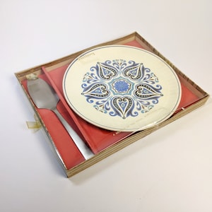 May include: A white ceramic cake plate with a blue and brown paisley design. The plate is in a cardboard box with a red lining and a silver cake server.