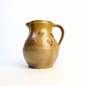 May include: A vintage ceramic pitcher in a warm, earthy tone. The pitcher has a rounded body, a curved handle, and a spout. It is decorated with embossed floral patterns, adding a rustic charm. This piece is ideal for serving beverages or as a decorative item.