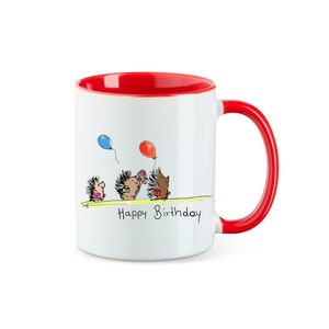 Könnte beinhalten: Weiße Keramiktasse mit rotem Rand und Henkel. Die Tasse zeigt eine Comic-Illustration von drei Igeln mit Ballons und dem Text "Happy Birthday".